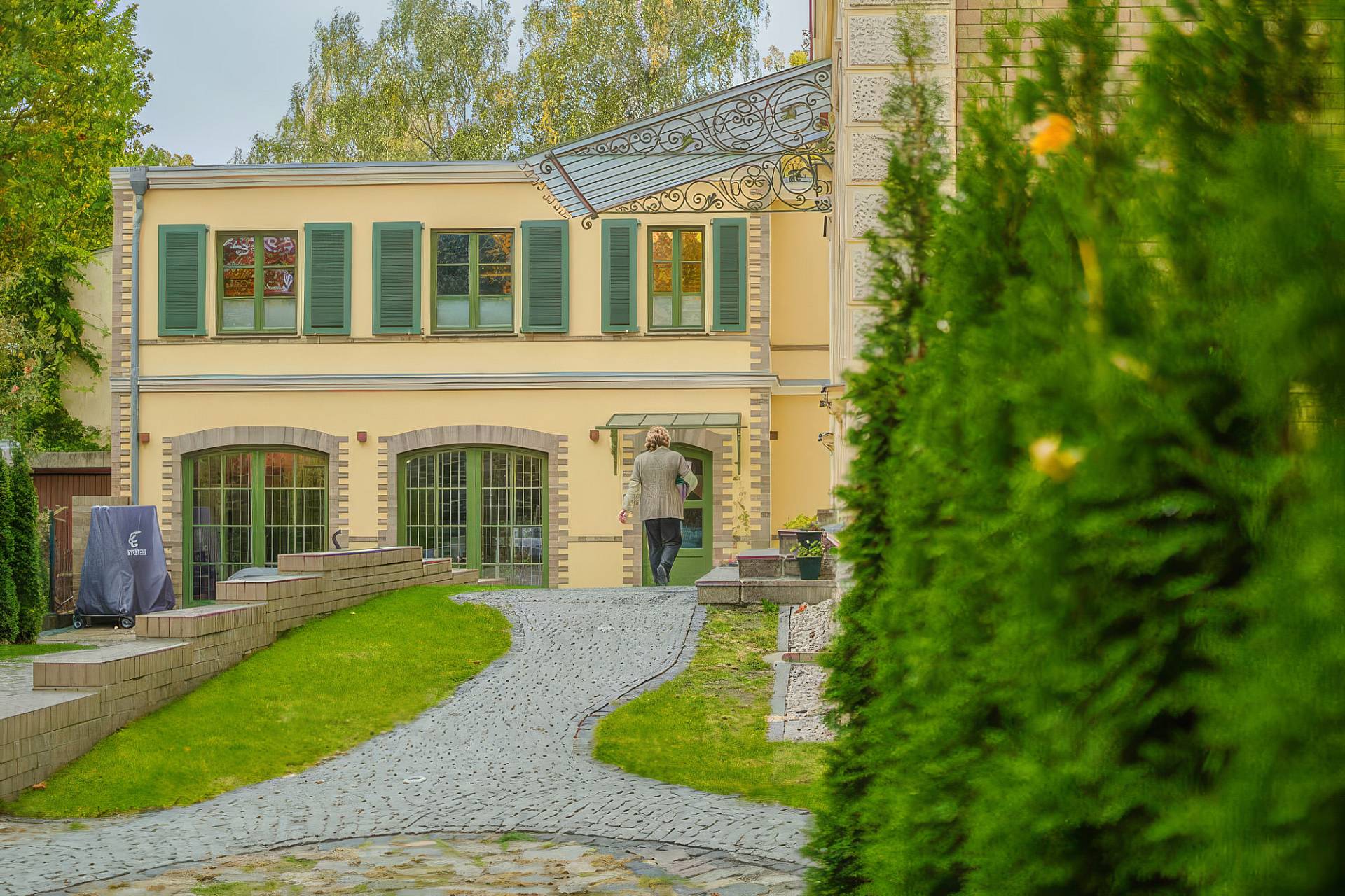 Gepflasterter Weg führt durch den Garten zum hellen Institutsgebäude mit grünen Fensterläden.