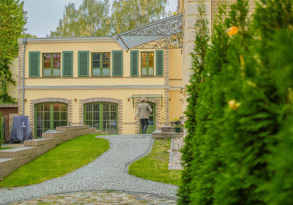Gepflasterter Weg führt durch den Garten zum hellen Institutsgebäude mit grünen Fensterläden.