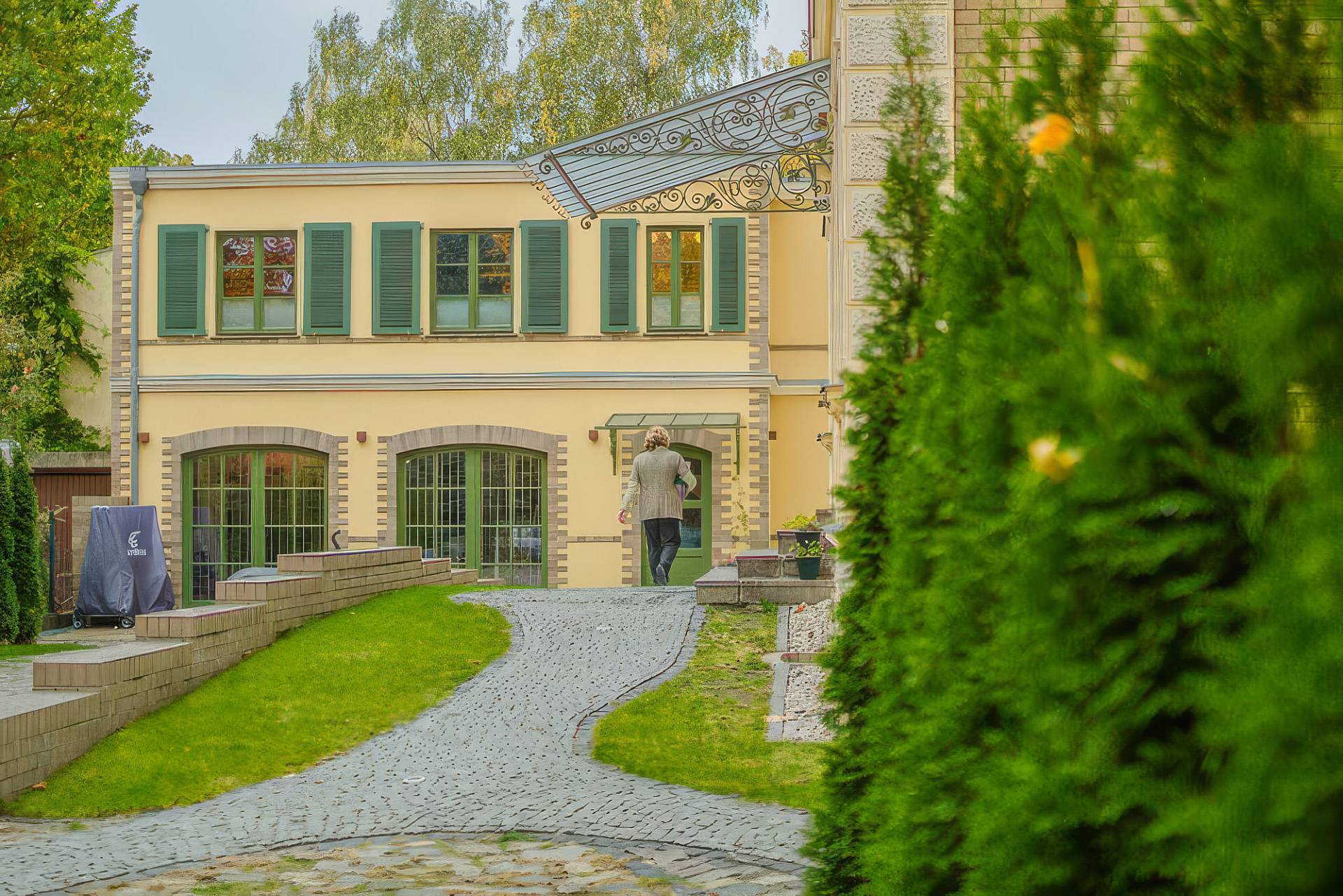 Gepflasterter Weg führt durch den Garten zum hellen Institutsgebäude mit grünen Fensterläden.