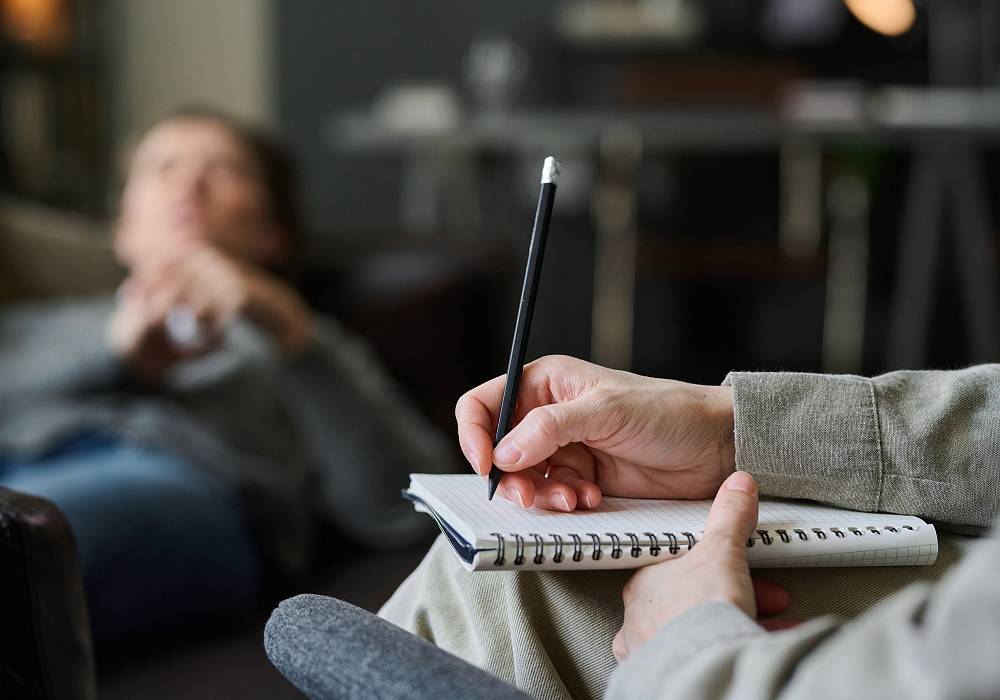 Therapeutin macht Notizen während einer Sitzung, im Hintergrund liegt eine Person entspannt auf dem Sofa.