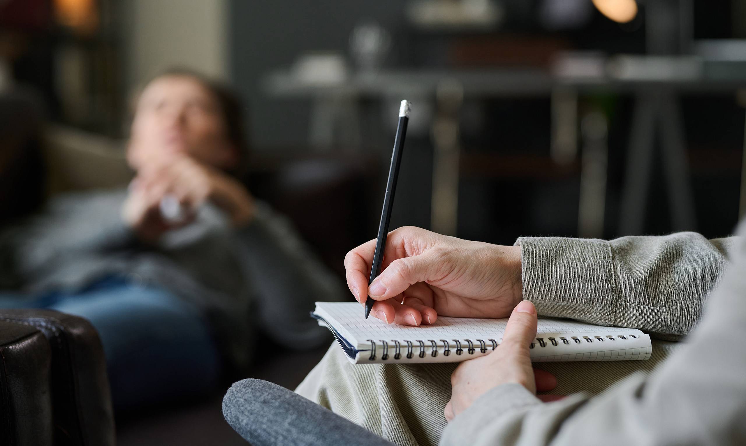 Therapeutin macht Notizen während einer Sitzung, im Hintergrund liegt eine Person entspannt auf dem Sofa.